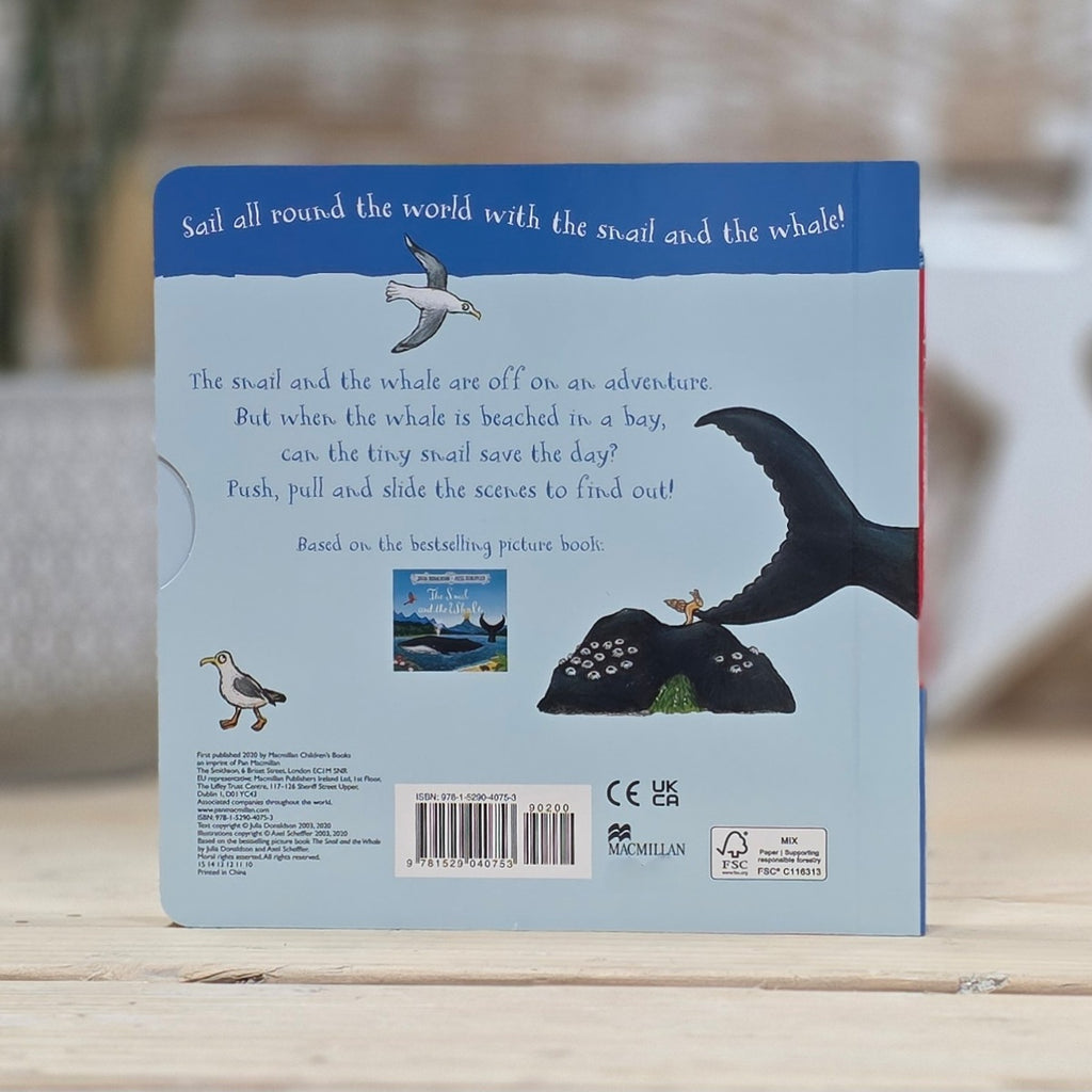 The back cover of a children's book titled 'Sail all around the world with the snail and the whale!' resting on a light wooden surface with a blurred outdoor background.