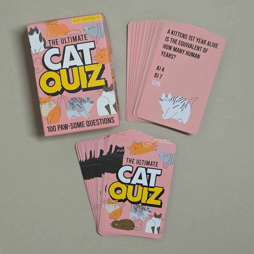 The Ultimate Cat Quiz card deck shown with its pink illustrated box and several fanned cards on a neutral gray surface. The cards feature pink backgrounds with black, yellow, white, and orange cat-themed graphics and printed quiz questions.