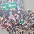 Boxed jigsaw puzzle titled "The World of Hercule Poirot" shown open with many puzzle pieces scattered around it on a light grey surface. The box features a vintage illustrated scene with people in a living room, and a green badge says "1000 pieces to solve the clues.