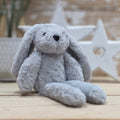 A light grey soft fuzzy plush bunny toy, with embroidered black eyes and nose, on a wooden surface with a blurred star-shaped object in the background