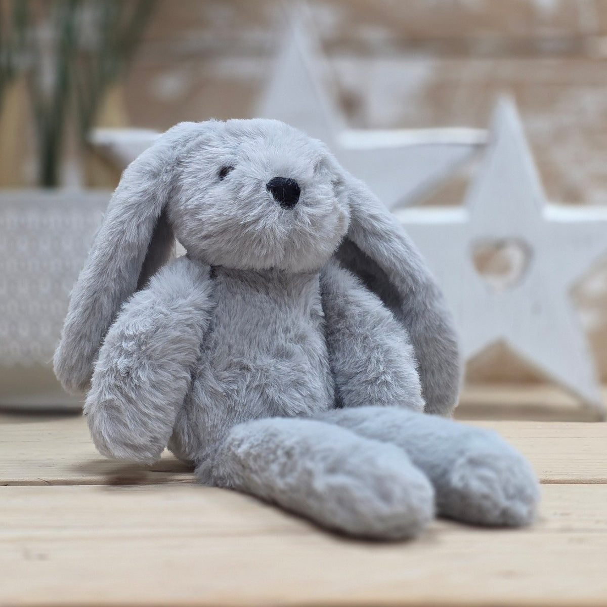 A light grey soft fuzzy plush bunny toy, with embroidered black eyes and nose, on a wooden surface with a blurred star-shaped object in the background