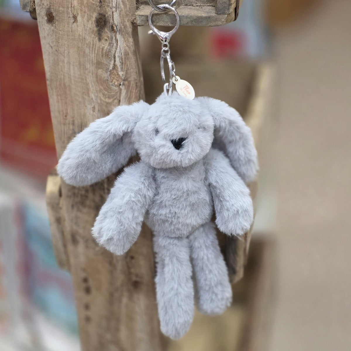 A light grey plush bunny keychain hanging from a rustic wooden post. The keychain features a soft, fuzzy grey bunny with embroidered black eyes and nose, attached to a silver metal key ring and a small round tag.