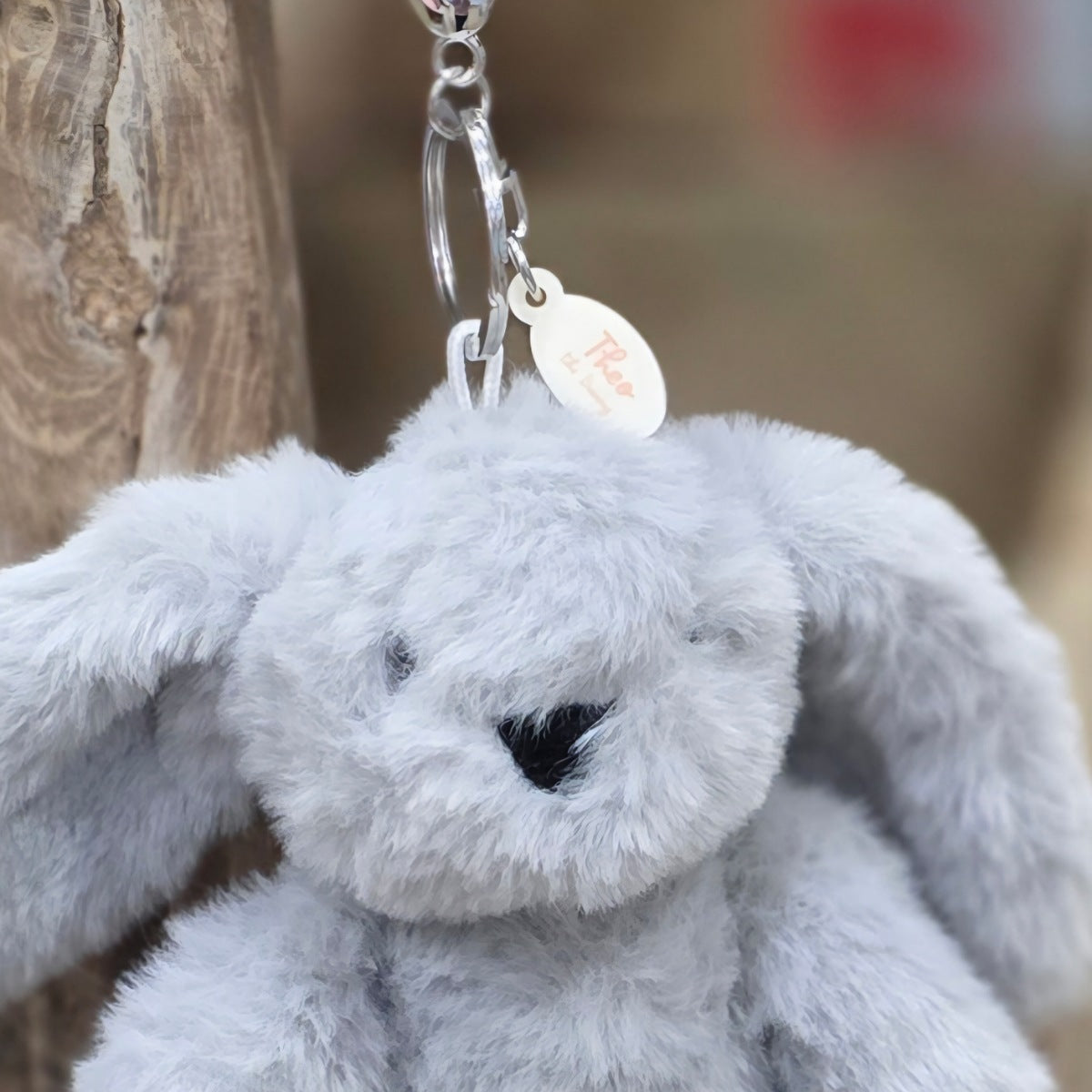 Grey plush bunny keychain with a tag against a blurred natural background