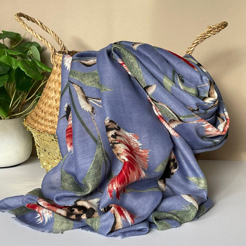 A denim blue scarf with an all over thistle print in olive, pink, red, white and black, draped over a woven straw basket next to a potted green plant.
