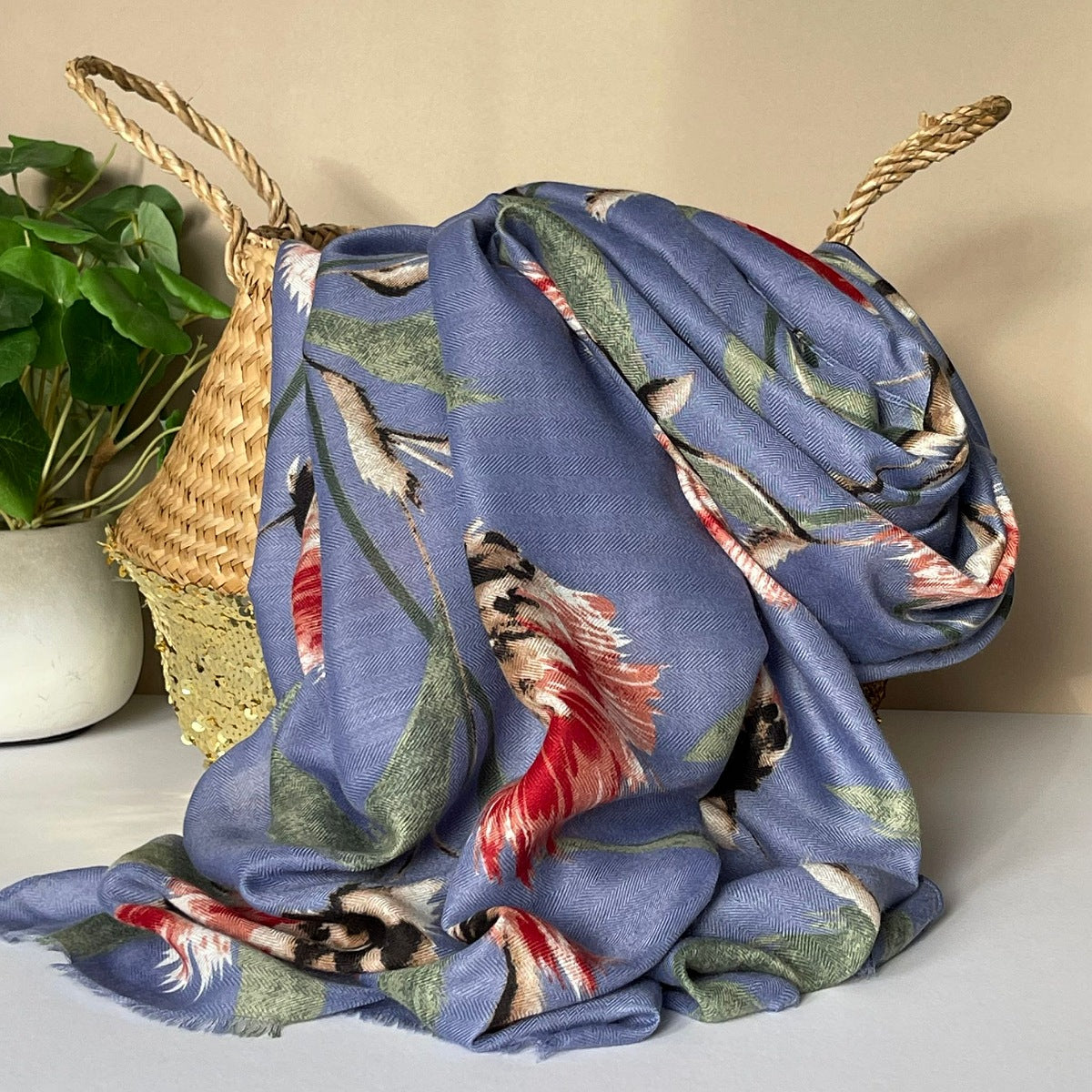 A denim blue scarf with an all over thistle print in olive, pink, red, white and black, draped over a woven straw basket next to a potted green plant.
