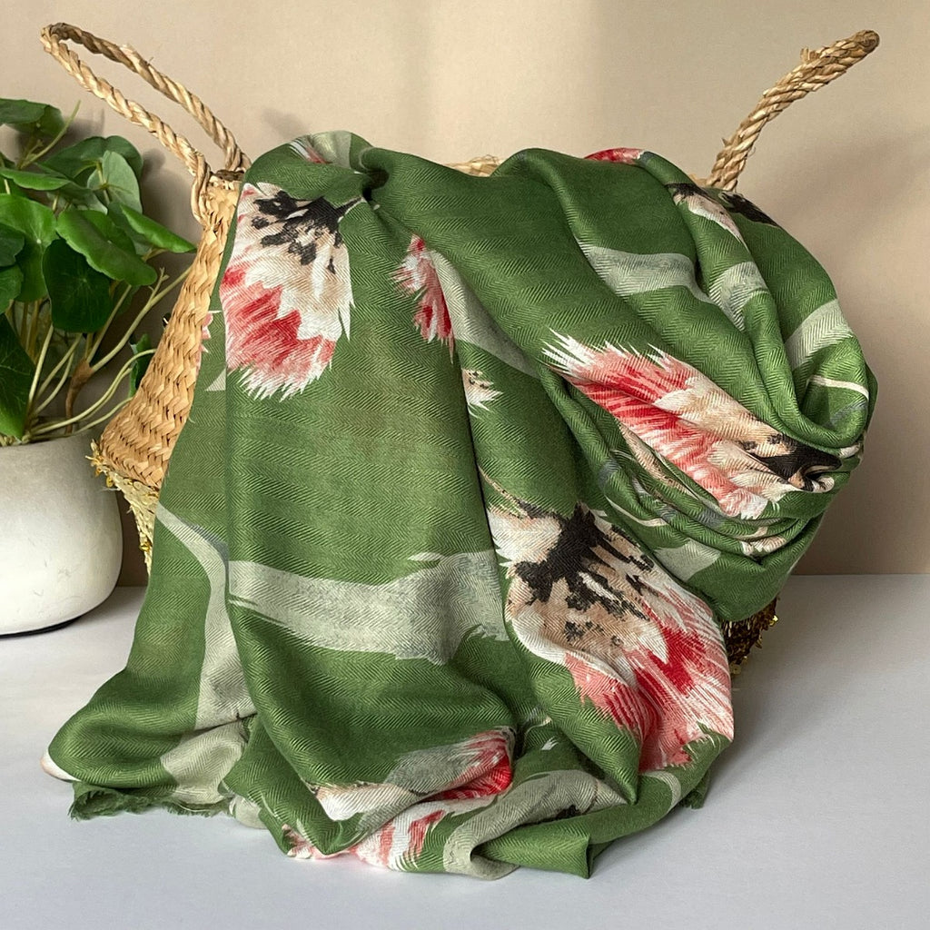 A green scarf with an all over thistle print in olive, pink, red, white and black, draped over a woven straw basket next to a potted green plant.
