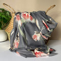 A grey scarf with an all over thistle print in olive, pink, red, white and black, draped over a woven straw basket next to a potted green plant.
