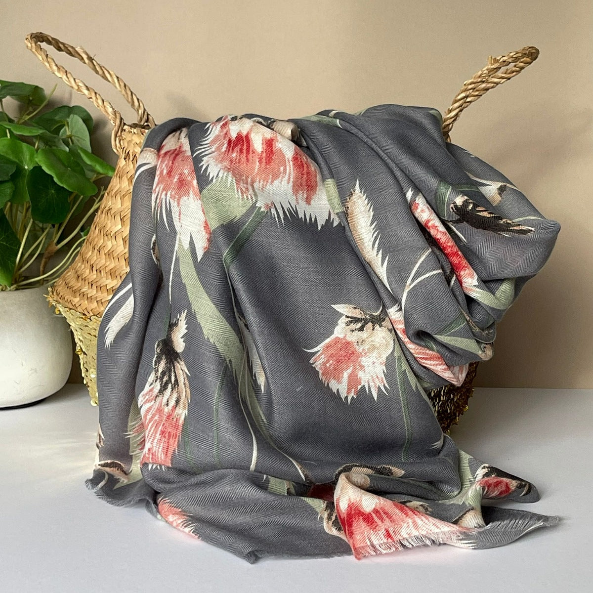 A grey scarf with an all over thistle print in olive, pink, red, white and black, draped over a woven straw basket next to a potted green plant.
