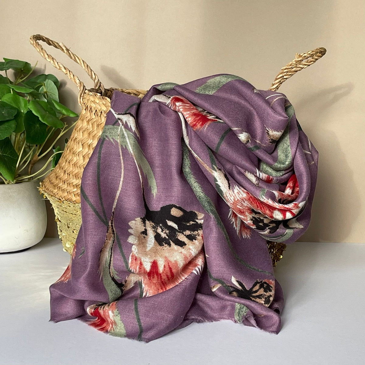 A purple scarf with an all over thistle print in olive, pink, red, white and black, draped over a woven straw basket next to a potted green plant.
