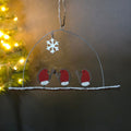 A hanging Christmas ornament shaped like three red-breasted birds on a branch under a wire dome, with a white snowflake and string hanger, photographed in front of a softly blurred Christmas tree with warm lights.