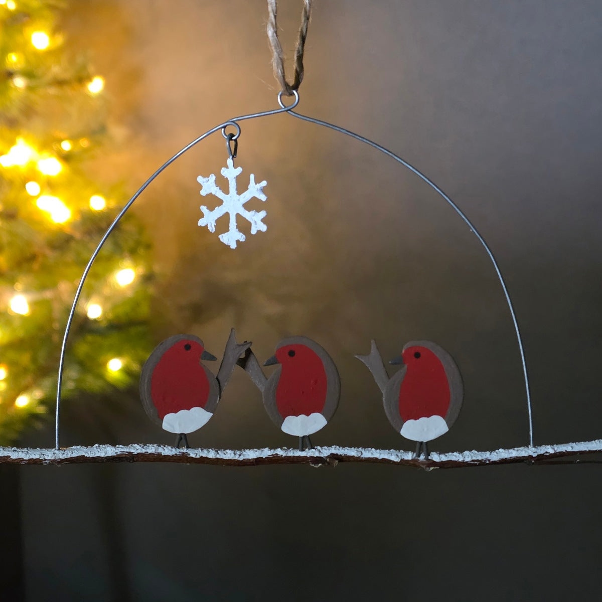A winter-themed metal ornament featuring three red-breasted robins under a wire dome with a hanging snowflake, displayed against a softly blurred Christmas tree and warm background lights.
