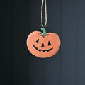 A metal hand-painted terracotta pumpkin ornament with a carved jack-o'-lantern face and a green stem, hanging from a natural jute twine string against a dark grae panelled background.