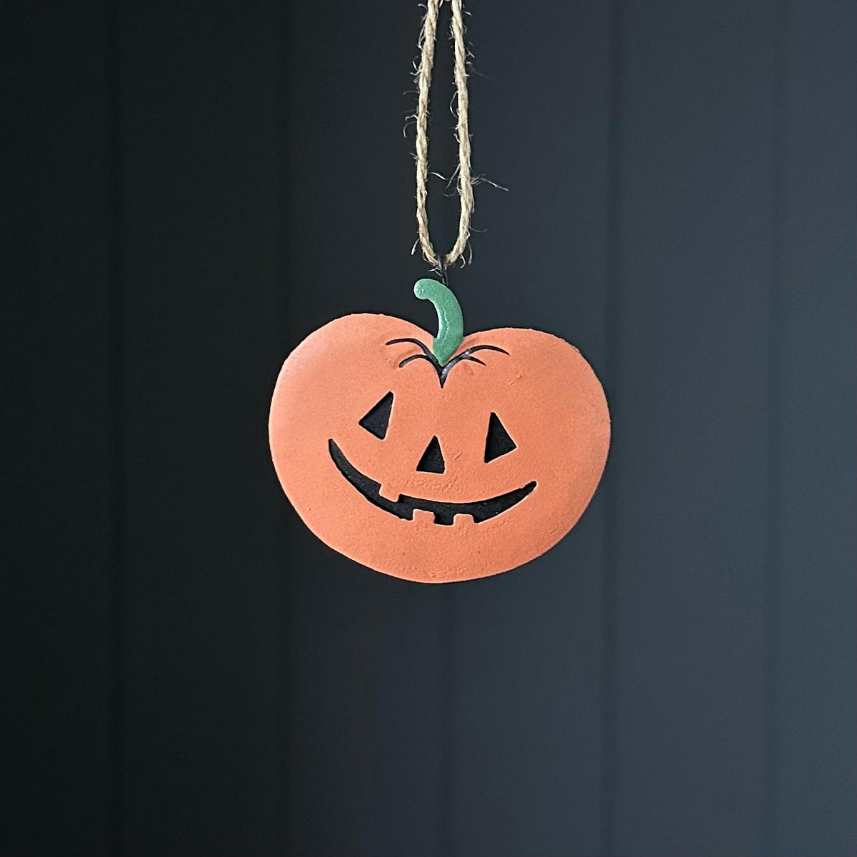 A metal hand-painted terracotta pumpkin ornament with a carved jack-o'-lantern face and a green stem, hanging from a natural jute twine string against a dark grae panelled background.