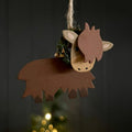 A rustic metal highland cow ornament hanging from a jute string against a dark background with a softly blurred Christmas tree and warm lights below.