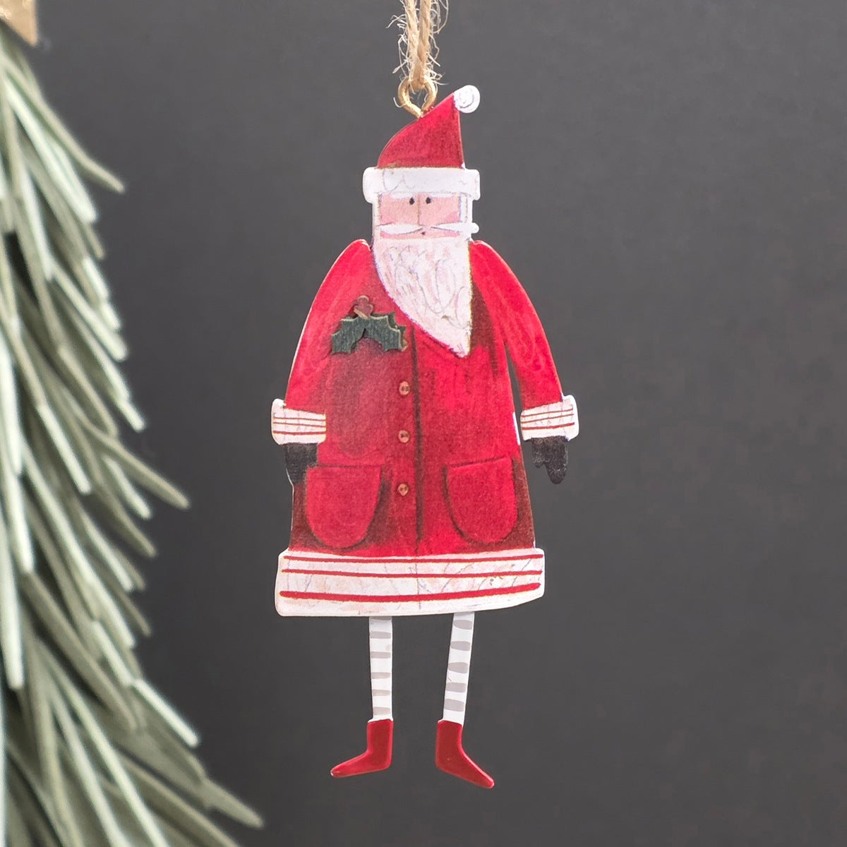 Handcrafted metal Santa Claus hanging ornament with red coat, white beard and trim, striped legs, and a gold hanging loop against a dark grey background with blurred pine needles on the left.