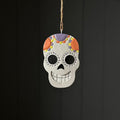 A hand-painted Day of the Dead sugar skull ornament hanging from a jute twine string against a dark grey panelled background.