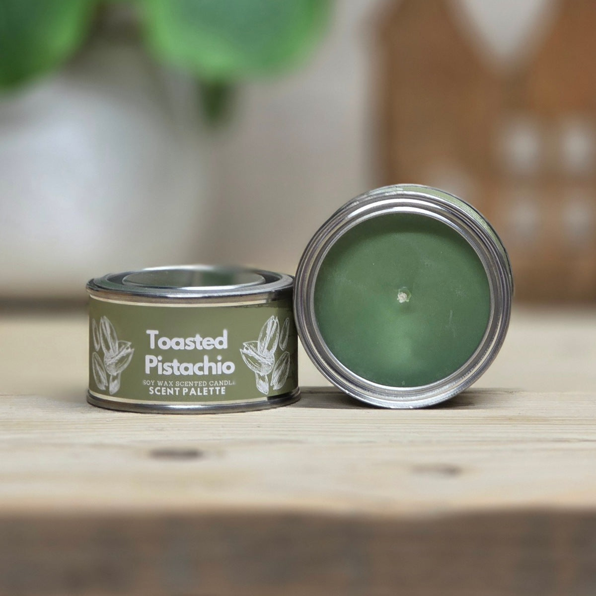 A small round metal tin candle labeled 'Toasted Pistachio' sits on a wooden surface with its lid leaning against it. The candle wax is visible inside, and the background is softly blurred with a plant and warm decor.