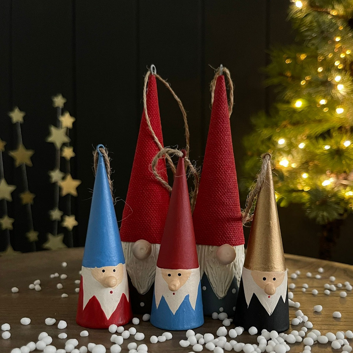 Five painted Christmas gnome ornaments with tall pointed hats in blue, red, dark blue, and gold, arranged on a wooden surface with white decorative beads and a softly blurred Christmas tree in the background.