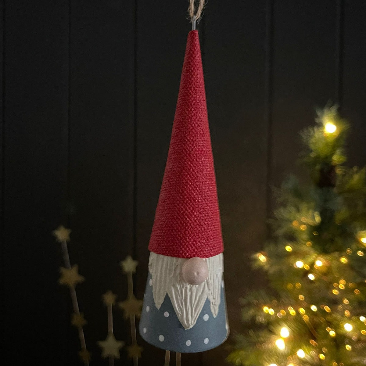 A hanging gnome ornament with a tall red burlap-textured hat, white beard, and blue polka-dotted base, shown against a dark panelled background with blurred warm lights and a Christmas tree on the right.