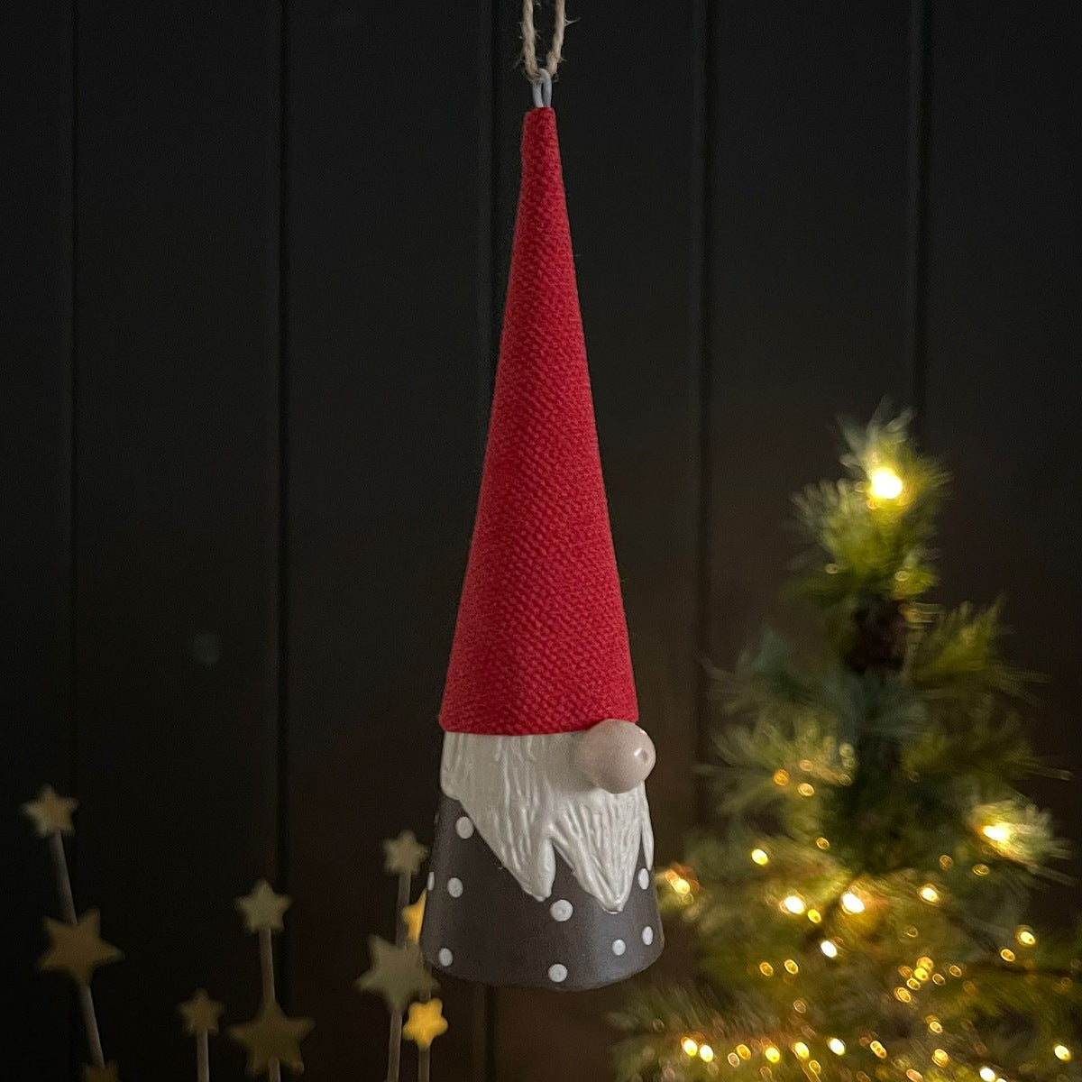 A hanging Christmas gnome ornament with a textured red hat, white beard, and gray body with white polka dots, shown in front of a dark panelled wall and a softly lit Christmas tree.