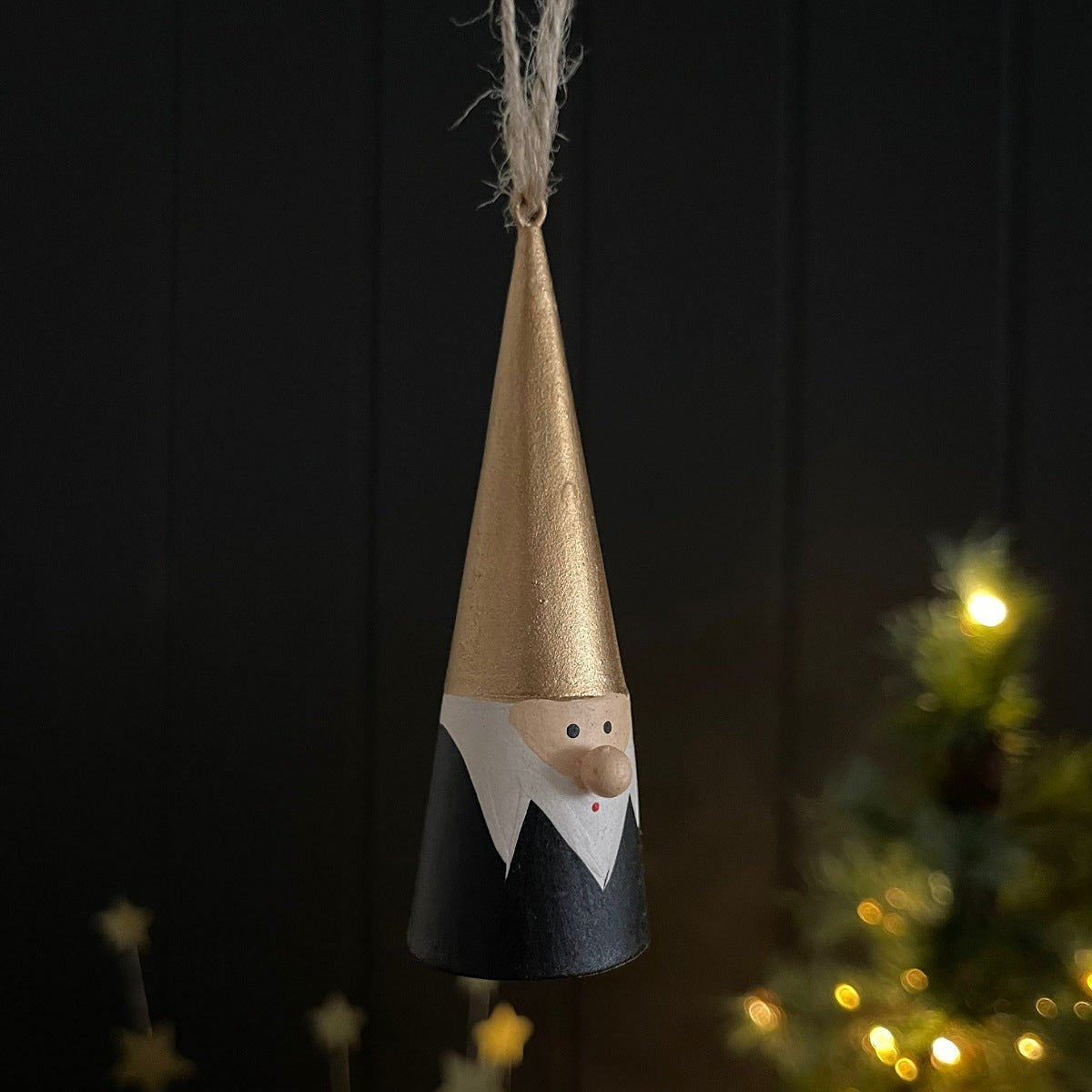 A hanging Christmas ornament shaped like a gnome, with a metallic gold pointed hat, black body, white collar, and a small beige face, suspended against a dark background with blurred warm lights in the distance.