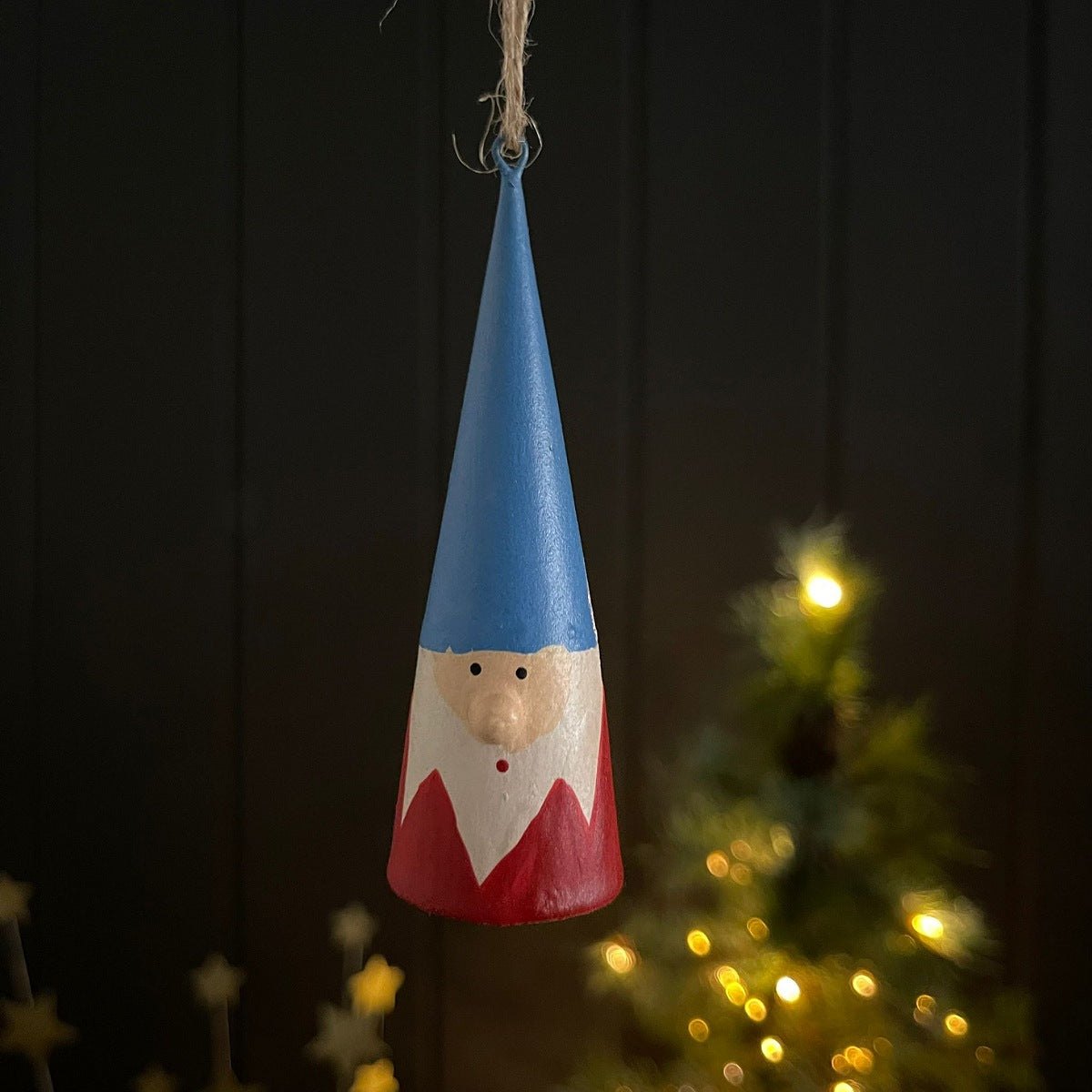 A hand-painted wooden gnome hanging ornament with a blue hat, red body, and white beard, suspended against a dark panelled background with a blurred Christmas tree and warm lights.