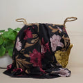 A black scarf with a large floral print in pink, burgundy, and beige, draped over a woven gold basket with rope handles, next to a green potted plant.