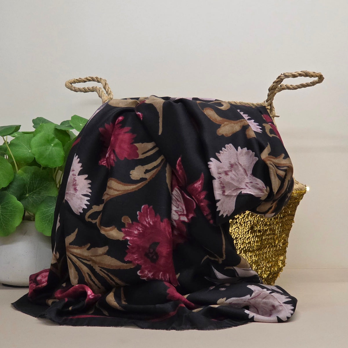 A black scarf with a large floral print in pink, burgundy, and beige, draped over a woven gold basket with rope handles, next to a green potted plant.