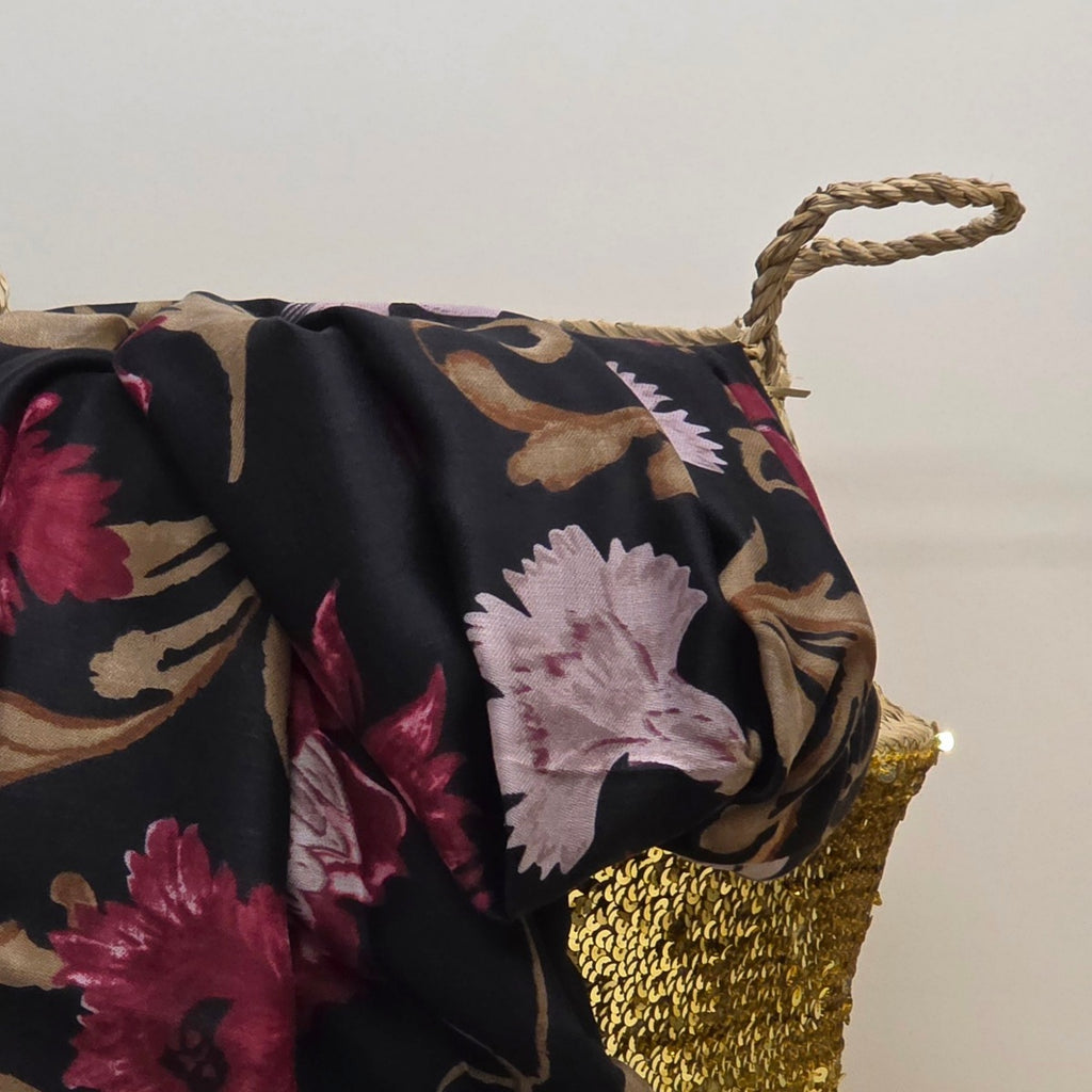A close-up of a draped black scarf with a floral pattern in pink, burgundy, and beige, draped over a woven basket with a gold woven base and rope handles.