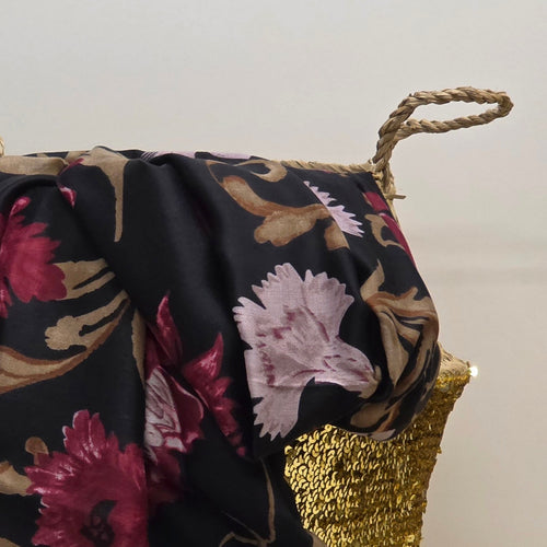 A close-up of a draped black scarf with a floral pattern in pink, burgundy, and beige, draped over a woven basket with a gold woven base and rope handles.