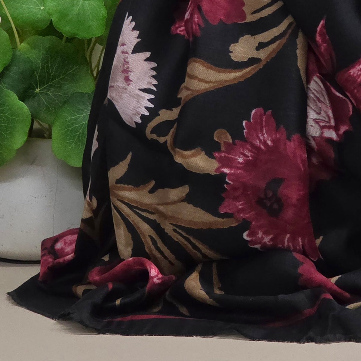 A black scarf with a large floral pattern in deep magenta and beige, draped next to a potted green plant.