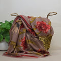 A draped floral scarf in dusty mauve with red, green, and yellow botanical patterns, styled over a gold sequined basket with rope handles, next to a green potted plant against a plain light background.