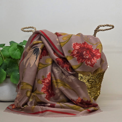 A draped floral scarf in dusty mauve with red, green, and yellow botanical patterns, styled over a gold sequined basket with rope handles, next to a green potted plant against a plain light background.