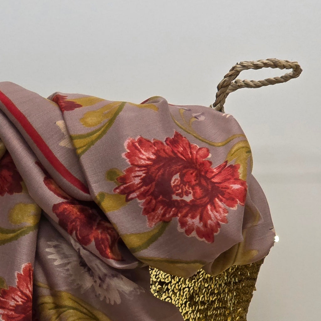 A draped floral scarf or shawl in muted mauve with large red flowers, olive-green leaves, and a red stripe, shown with a braided rope handle against a plain light background.