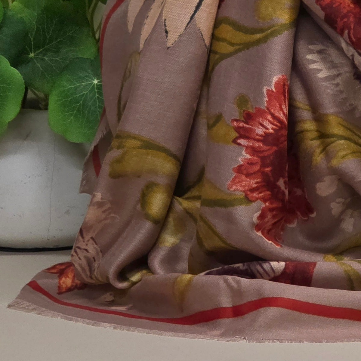 A draped silk scarf with a muted taupe base, featuring a large red floral motif, olive green leaves, and a thin red stripe along the fringed edge, styled next to a potted green plant.