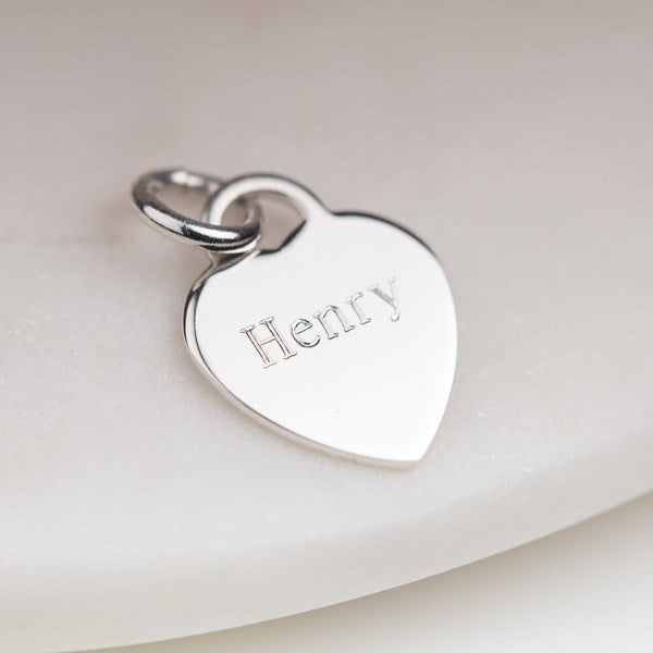 A silver heart-shaped pendant charm engraved with the name 'Henry', resting on a soft, light-colored surface.