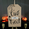 A wooden Halloween tag sign with 'Trick or Treat' written in black script, hanging in front of a dark paneled wall with glowing jack-o'-lanterns on a shelf behind it.