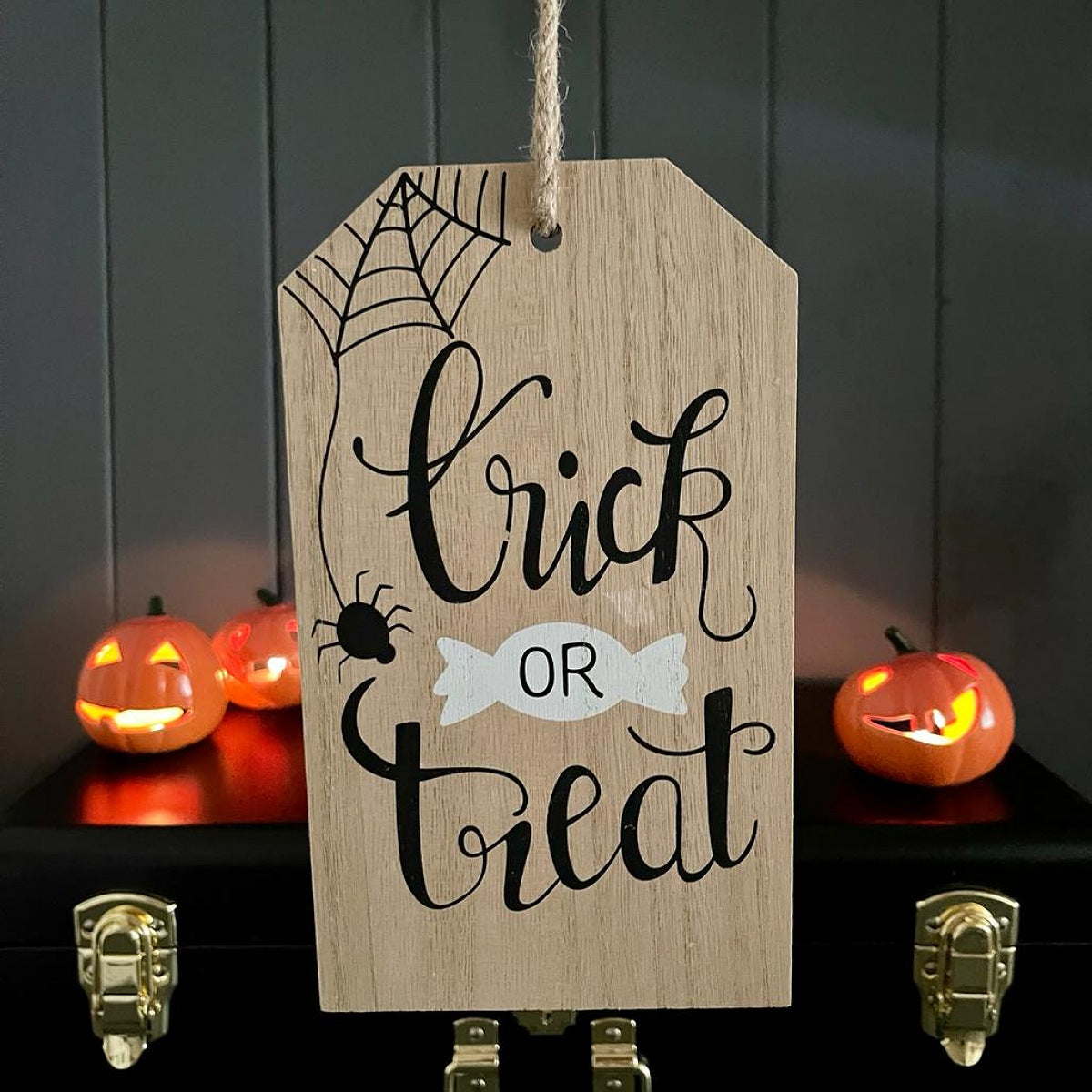 A wooden Halloween tag sign with 'Trick or Treat' written in black script, hanging in front of a dark paneled wall with glowing jack-o'-lanterns on a shelf behind it.