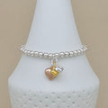 A silver beaded bracelet with two dangling heart charms, one rose gold and one silver, displayed on a white cone-shaped jewelry stand against a beige background.