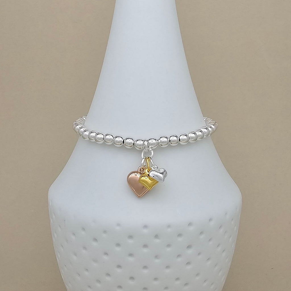 A silver beaded bracelet with two dangling heart charms, one rose gold and one silver, displayed on a white cone-shaped jewelry stand against a beige background.