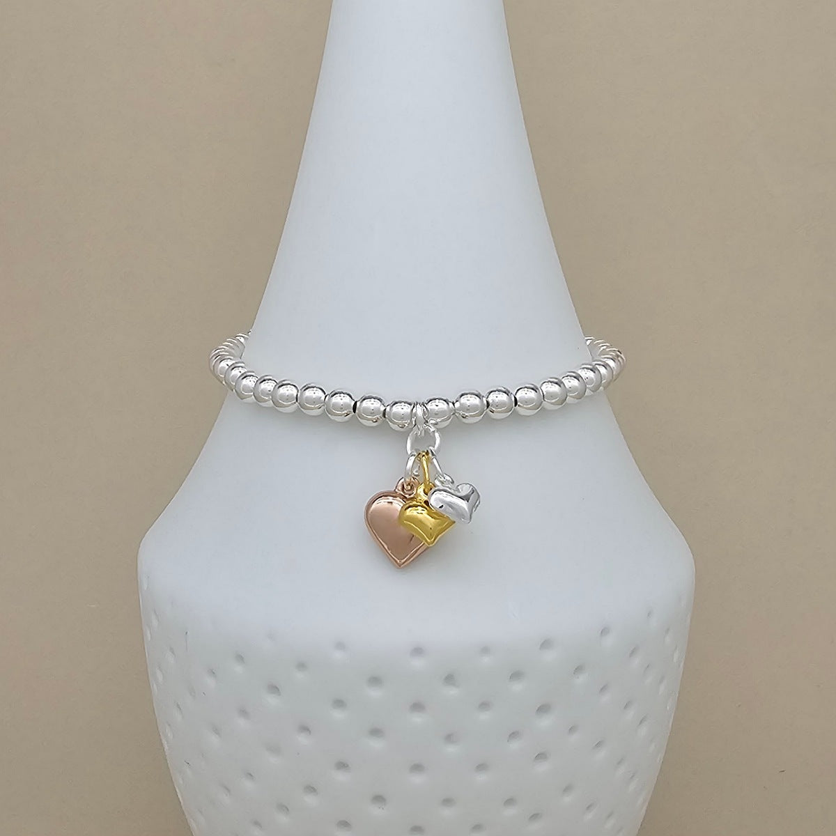 A silver beaded bracelet with two dangling heart charms, one rose gold and one silver, displayed on a white cone-shaped jewelry stand against a beige background.