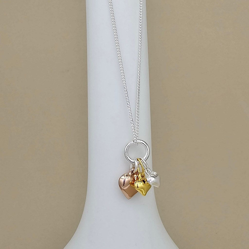 A silver chain necklace with three small heart charms in rose gold, yellow gold, and silver, displayed on a white jewelry bust against a beige background.