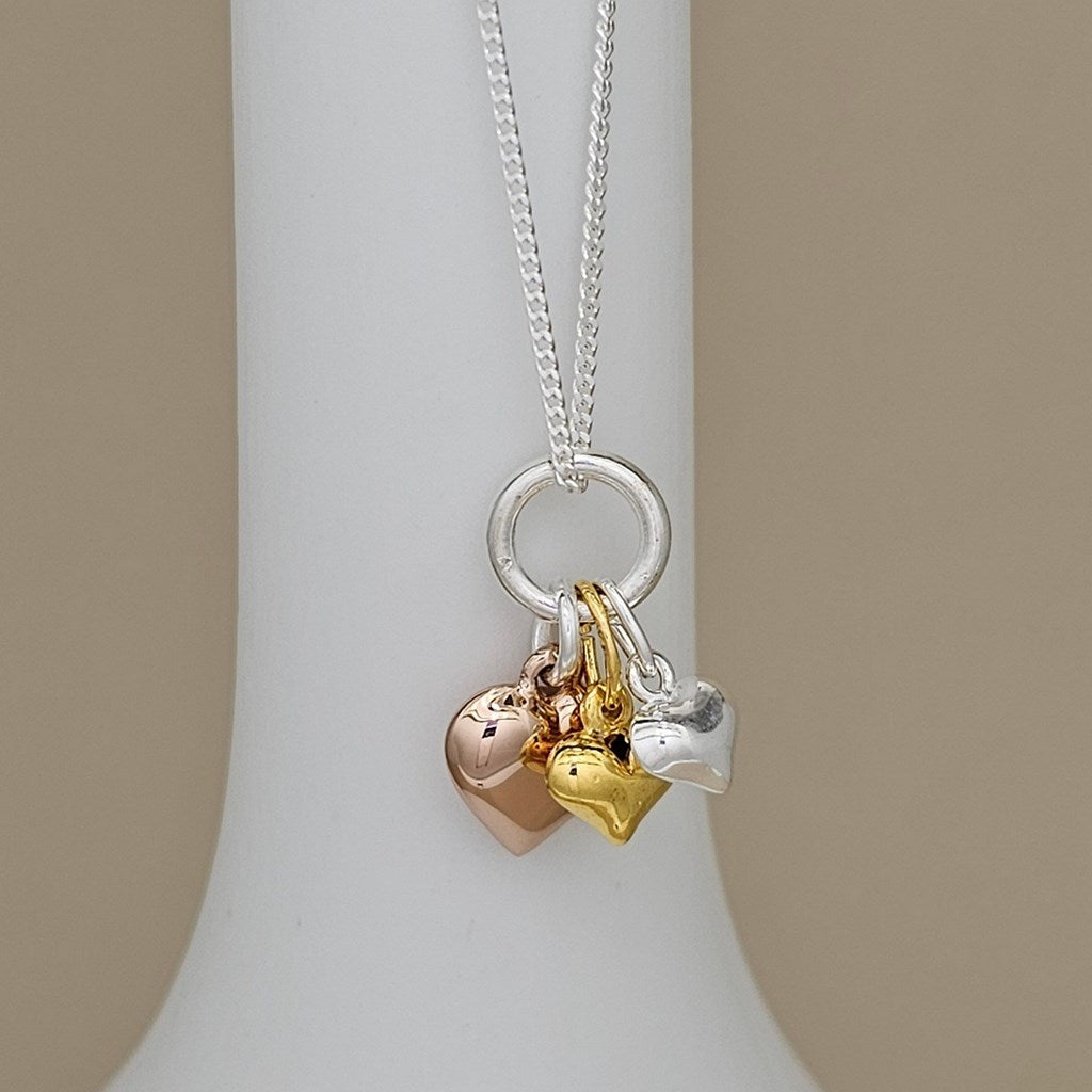 A silver chain necklace with a round ring and three dangling heart charms in rose gold, yellow gold, and silver, displayed on a white jewelry bust against a beige background.