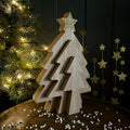 A wooden triple Christmas tree-shaped decorative object with a glittery gold star topper, displayed on a table with a blurred Christmas tree and hanging gold stars in the background.