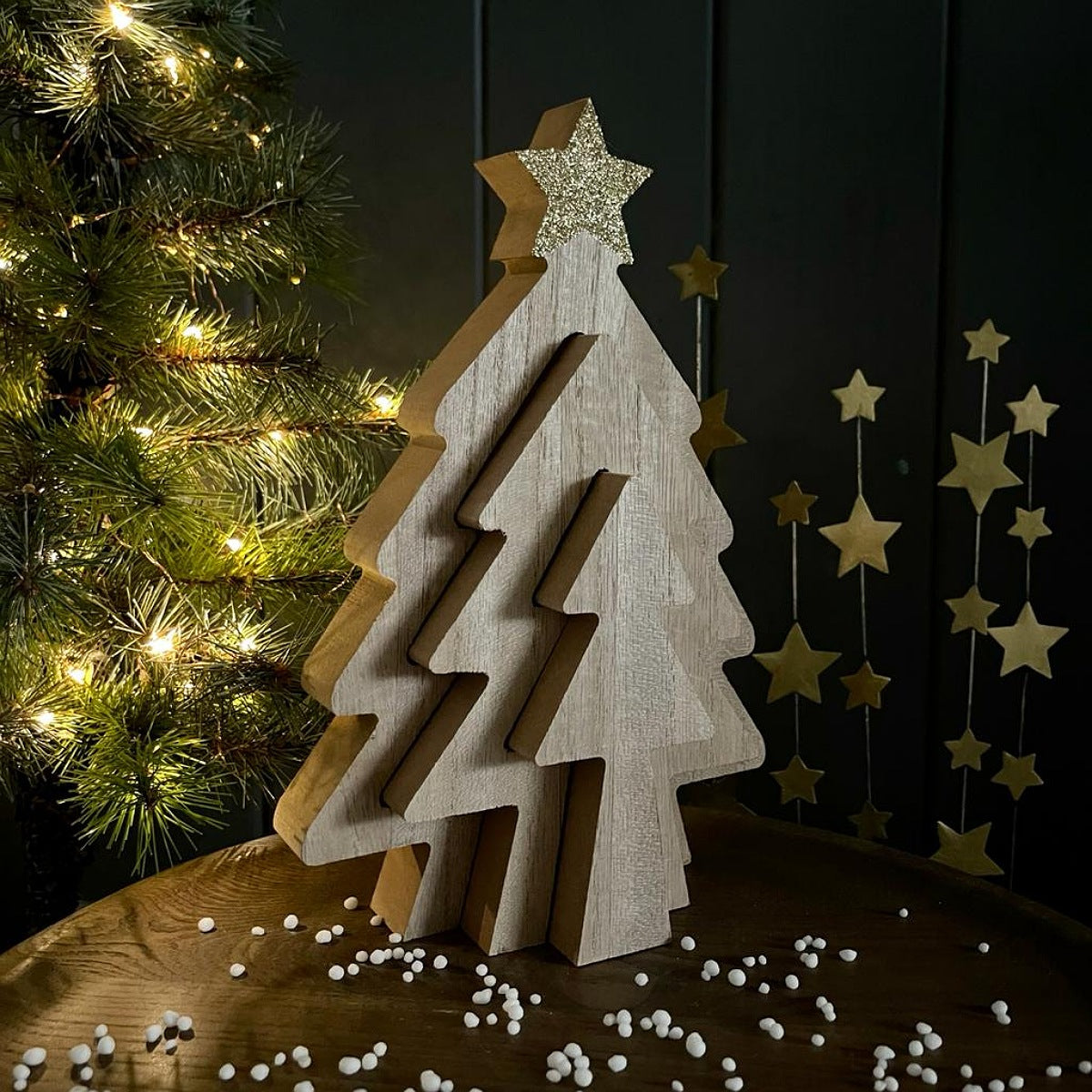 A wooden triple Christmas tree-shaped decorative object with a glittery gold star topper, displayed on a table with a blurred Christmas tree and hanging gold stars in the background.