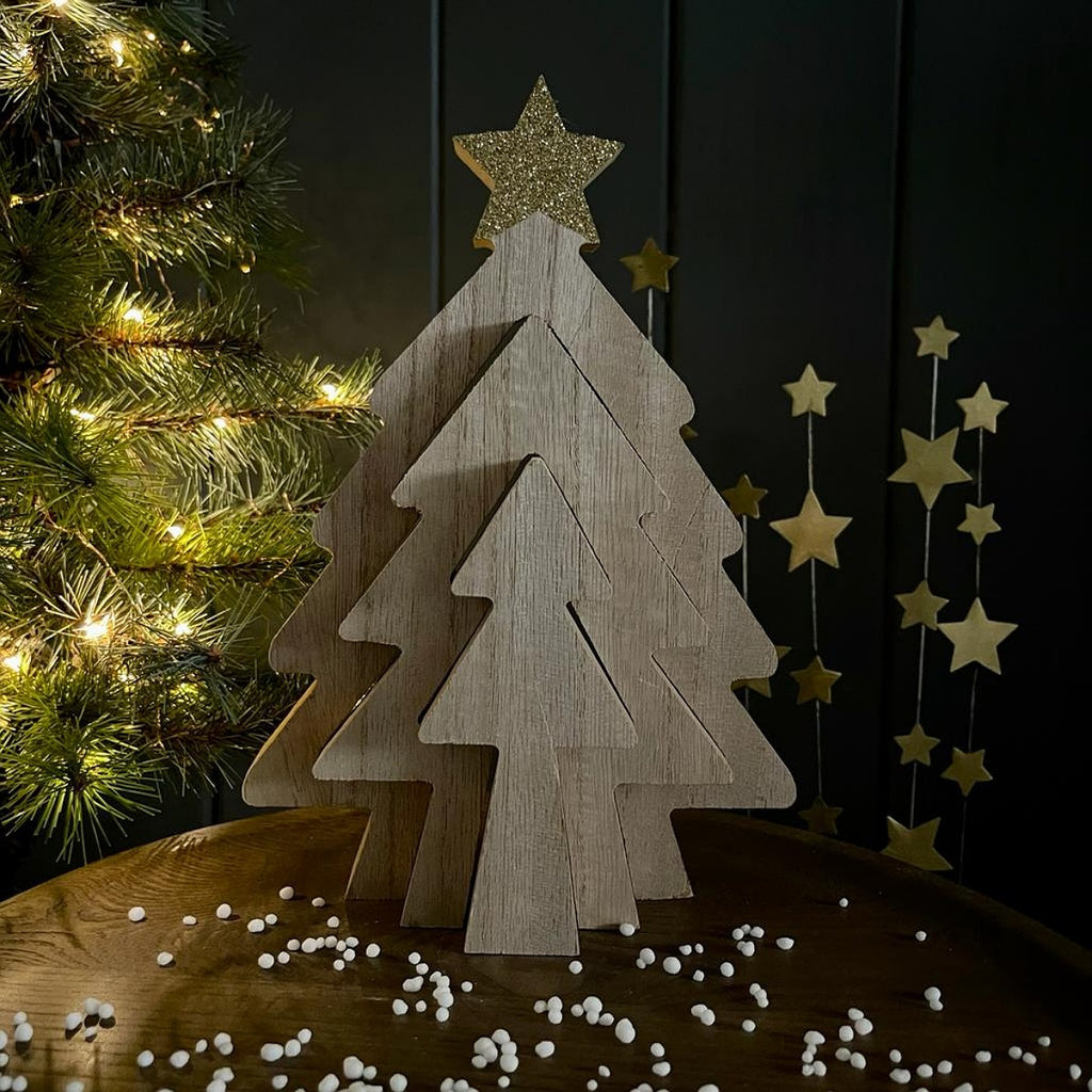 A wooden Christmas tree tabletop decoration made of layered triangular wood cutouts with a glittery gold star topper, displayed on a wooden surface with small white beads scattered around, a lit Christmas tree at left, and gold hanging star garlands against a dark background.