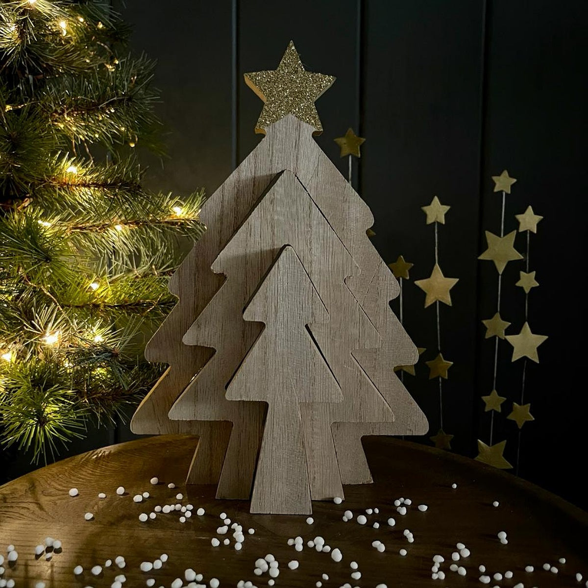 A wooden Christmas tree tabletop decoration made of layered triangular wood cutouts with a glittery gold star topper, displayed on a wooden surface with small white beads scattered around, a lit Christmas tree at left, and gold hanging star garlands against a dark background.
