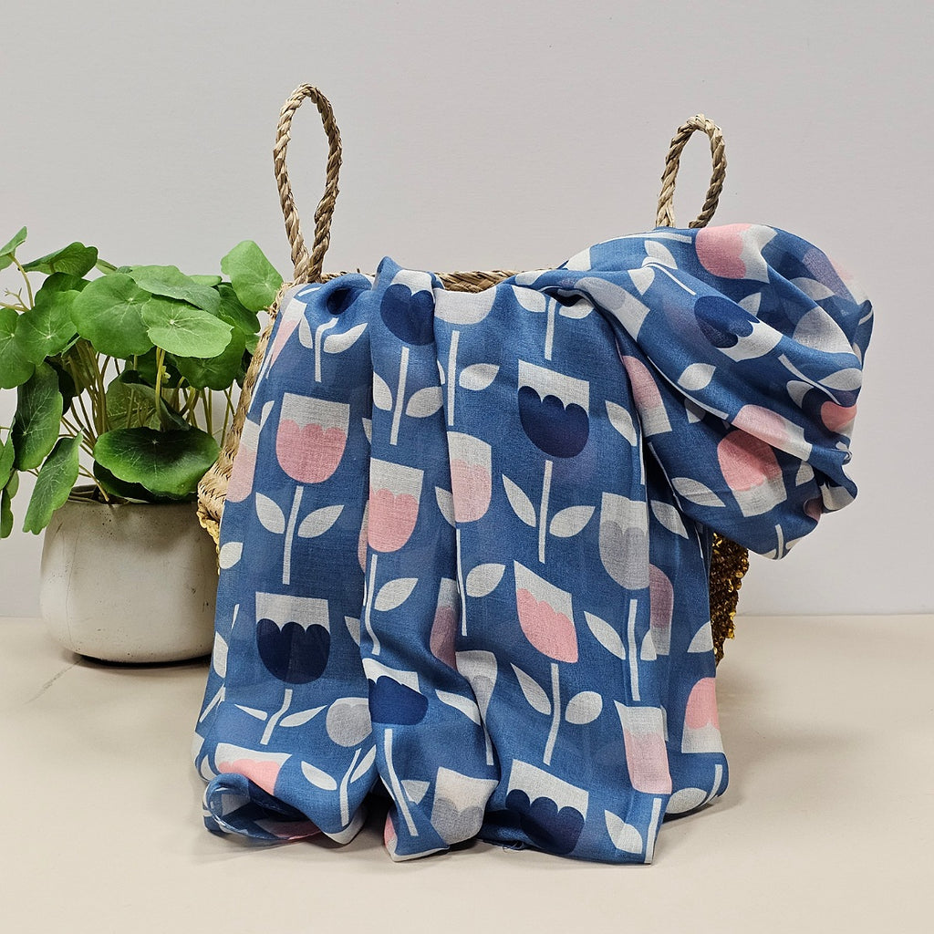 Blue scarf with tulip prints in pink, navy and grey draped over a wicker basket.  Styled on a light beige surface next to a potted green plant against a neutral light grey background.