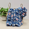 Blue scarf with tulip prints in pink, navy and grey draped over a wicker basket.  Styled on a light beige surface next to a potted green plant against a neutral light grey background.