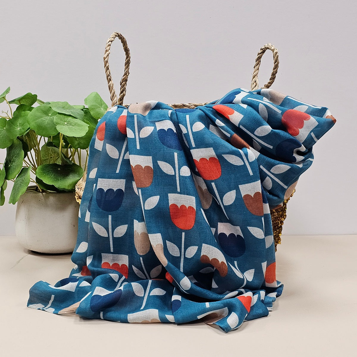 Teal blue scarf with tulip prints in red, navy, brown, and beige draped over a wicker basket.  Styled on a light beige surface next to a potted green plant against a neutral light grey background.